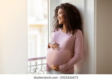 Radiant black pregnant woman gently cradling her belly while standing by window at home, beautiful african american expectant mother gazing outward thoughtfully, enjoying pregnancy time, copy space - Powered by Shutterstock