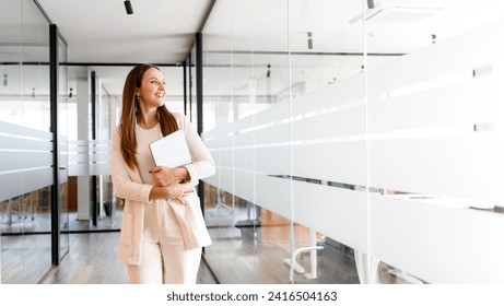 A radiant businesswoman holding a laptop looks aside with a satisfied expression standing in a brightly glass corridor, symbolizing ambition and forward-thinking in the corporate world - Powered by Shutterstock