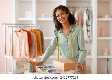 Radiant female fashion designer leaning on a desk with a package, color swatches, and glasses, ready to work in her bright, organized studio - Powered by Shutterstock