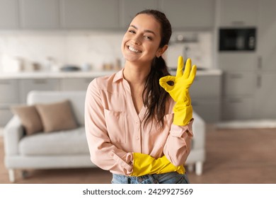 Radiant woman in pink blouse and cleaning gloves making an okay gesture, symbolizing satisfaction with her cleaning in bright kitchen, free space - Powered by Shutterstock