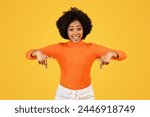 Radiant young woman with curly hair smiling broadly and pointing down with both hands, wearing a bright orange turtleneck and white pants against a striking yellow background