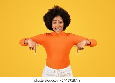 Radiant young woman with curly hair smiling broadly and pointing down with both hands, wearing a bright orange turtleneck and white pants against a striking yellow background - Powered by Shutterstock