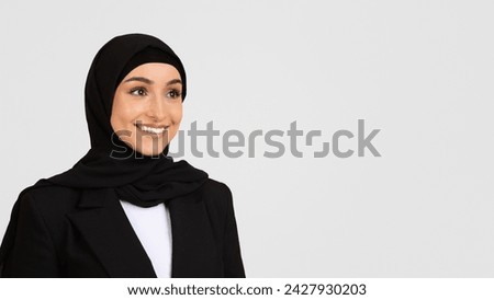 Radiant young woman wearing hijab and black blazer with beaming smile, standing against light grey background, embodying professionalism, free space