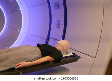 Radiation Proton Therapy. Brain Cancer Child Patient Lying On Positioning Bed With Fixing Mask On Her Head In Treatment Room.