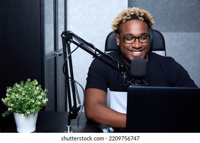 Radio DJ With Glasses Leads A Popular Program For Listeners. A Cheerful Black Man Podcaster At The Workplace With A Professional Microphone Sits In The Studio. 