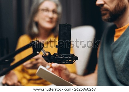 Radio host taking notes while interviewing a guest, using a professional microphone in a home studio setting. Focus on the microphone captures the essence of engaging conversation