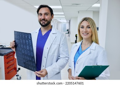 Radiographer With MRI Of Patient Head Is Standing In Corridor Of Hospital With Female Radiologic Technologist And At Looking At Camera. Team Working In Clinical Imaging