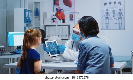 Radiologist Explaining X-ray Using Tablet In Medical Office And Nurse Working On Computer. Pediatrician Specialist With Protection Mask Providing Health Care Service Radiographic Treatment Examination