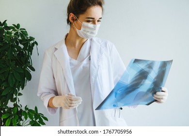 A Radiologist In A Medical Mask And Gloves Radiates An X-ray Image Of The Patient's Lungs. The Girl Doctor Works On The Front Line. The Doctor Is Holding An X-ray Of His Lungs.