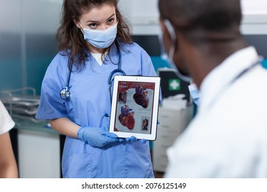 Radiologist Nurse With Protective Face Mask To Prevent Infection With Coronavirus Holding Tablet With Heart Expertise On Screen During Clinical Appointment. African American Doctor In Hospital Office