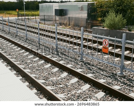 Rail corridor bordered by dense trees and shrubs includes gravel ballast, metal rails, and chain-link fence with multiple warning signs. Metal-sided utility structure with window AC units nearby. USA