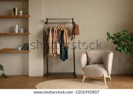 Rail rack with a capsule wardrobe in the living room against a beige wall. Trendy outfits in autumn colors: burgundy, beige, khaki, green, brown.
