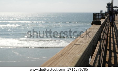 Railings of old wooden pier, people walking on waterfront boardwalk, Oceanside beach atmosphere, California coast USA. Defocused seascape, pacificocean water waves. Los Angeles summertime vacations.