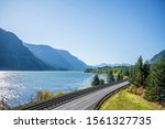 A railroad track next to a highway road with lane dividing opposite traffic runs along the scenic Columbia River bank with green trees and rocky mountains in Columbia Gorge area