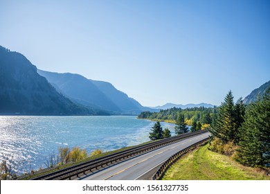 A Railroad Track Next To A Highway Road With Lane Dividing Opposite Traffic Runs Along The Scenic Columbia River Bank With Green Trees And Rocky Mountains In Columbia Gorge Area