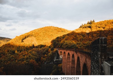 Railway bridge with arches spans across a valley filled with autumn foliage, with sunlit hills and a cloudy sky in the background.

 - Powered by Shutterstock