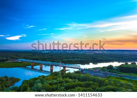 Railway bridge is reflected over the calm White River, surrounded by a green lush forest on the outskirts of the city under the evening blue sky at dusk. Sunset over Ufa, Bashkortostan, Russia.