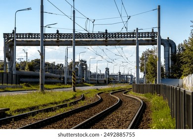 Railway tracks curve under industrial pipelines and overhead wires in an urban setting. The image highlights transportation and industrial infrastructure beneath a clear blue sky. - Powered by Shutterstock
