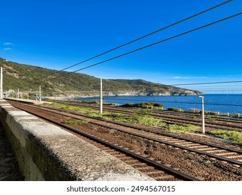 Railway tracks running alongside a stunning coastal landscape with lush green hills and the sparkling blue sea under a clear sky, creating a scenic and tranquil view. - Powered by Shutterstock
