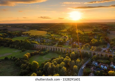 The railway viaduct at Chappel and Wakes Colne in Essex, England the sun a gold ball just above the horizon casting rays light and shadows across the landscape making the tops of the trees glow - Shutterstock ID 1528291601