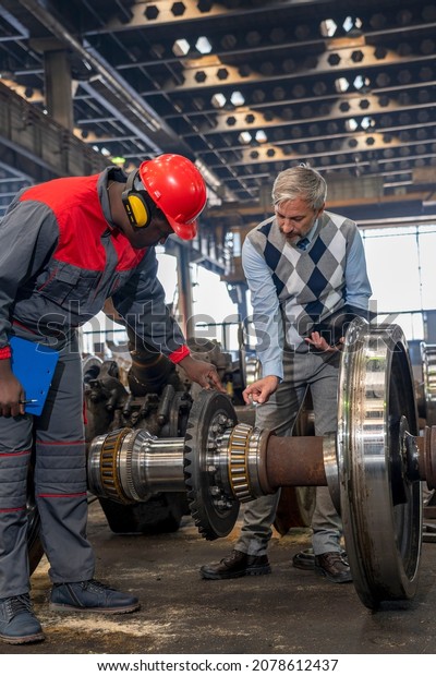 Railway Wheel Set Maintenance At Train Factory.\
Production Manager Talking To African American Worker In Protective\
Workwear Next To Train Wheels. Area For Maintenance, Repair And\
Service Of Trains.