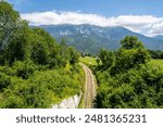 A railway winds through a lush valley, with Julian Alps in the backdrop. Enjoy a scenic train ride through Slovenias stunning countryside on a sunny summer day.