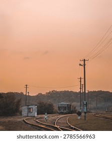 A railway yard at sunset with multiple train tracks curving and converging toward the horizon. A small brick building stands near the tracks, while a freight train car is visible further down the line. Utility poles with power lines run alongside the tracks, and dense trees form the background. The warm orange sky creates a calm and atmospheric industrial scene.