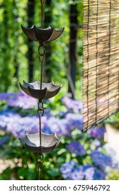 Rain Chains And A Bamboo Shade Before A Japanese Garden Where Blue Hydrangeas Are In Bloom.