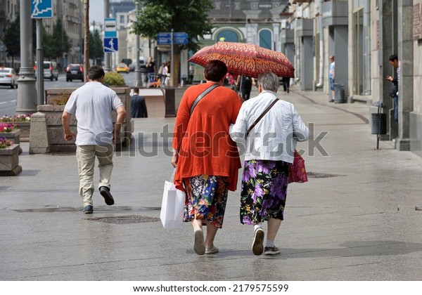 Rain in a city, two old women with one umbrella\
walk on a street on people background. Rainy weather, life of\
elderly people in summer