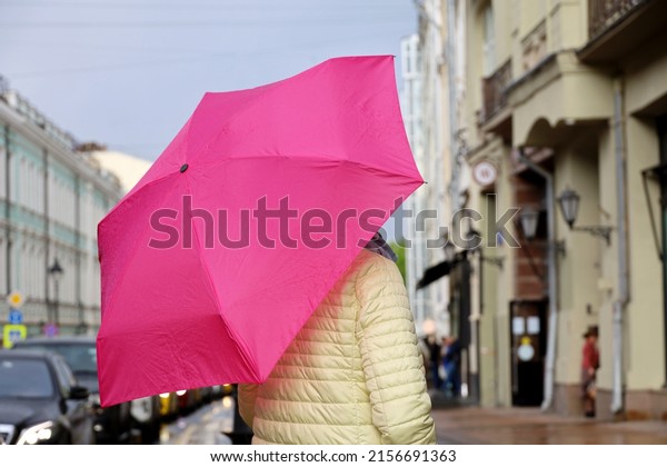 Rain\
in a city, woman standing with pink umbrella on city street on\
background of residential buildings and parked\
cars