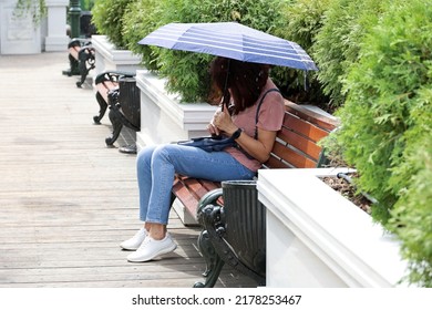 Rain In A City, Woman With Umbrella Sitting On Bench And Using Smartphone. Rainy Weather In City, Summer Storm