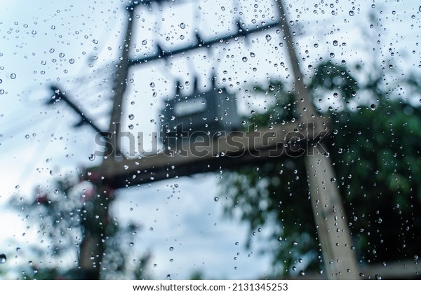 Rain\
droplets on surface of car glass with blurred electric pole with\
transformer and fuse background through window glass of car covered\
by raindrops. Wet windscreen shot from inside\
car.