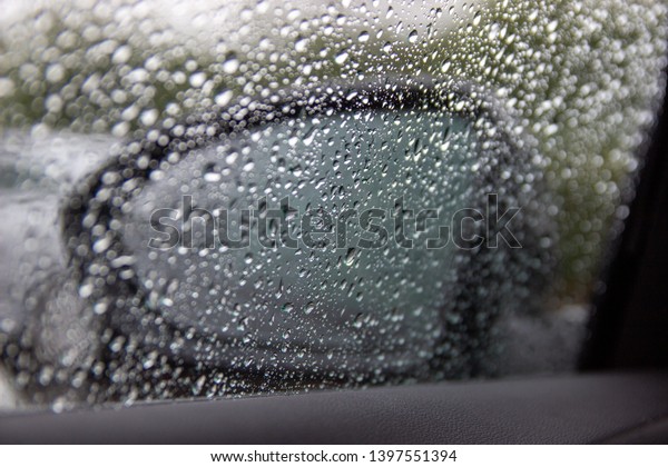 Rain drops on a window with a car wing mirror\
visible behind