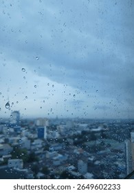 Rain drops on a window overlooking a blurry cityscape under a cloudy sky, evoking a calm, moody atmosphere.
