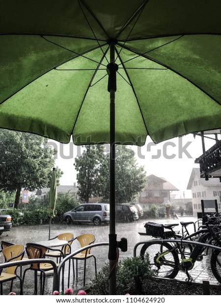 A rain shower on a summer day. View out of a green\
parasol on the rain. Empty tables, chairs, cars, bikes,  trees and\
a street. All wet with visible raindrops falling from the grey but\
bright sky.