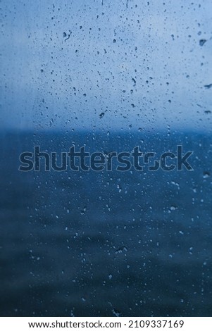 Rain and snow drops texture on the scratched window glass background. Oily surface. Deep blue sea behind. Selective focus.