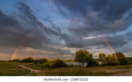 Rainbow Against A Dramatic Sky. Picturesque Rural Landscape. The Trees In The Foreground Are Illuminated By The Rays Of The Setting Sun.