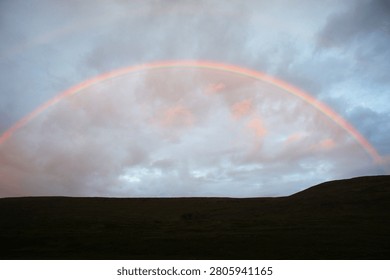 Rainbow arching over a dark hilly landscape under a cloudy sky. Natural meteorological weather phenomenon. Vibrant spectrum of light after rain on a gloomy day. Hope and inspiration.
