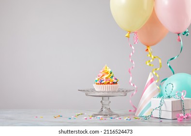 Rainbow Birthday cupcake on a stand with presents, hats and colorful balloons over light grey background. Scene from a birthday party!