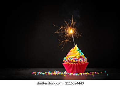 Rainbow Birthday cupcake with a sparkler and colorful sprinkles over a dark background. A magical Birthday celebration.