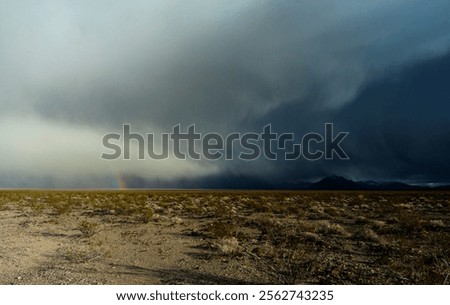 A rainbow breaks through storm clouds over the Nevada desert near Death Valley, revealing a dramatic landscape of rugged terrain, distant mountains, and moody skies.