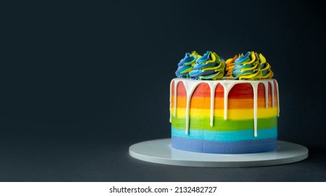 Rainbow cake with whipped cream top on the dark grey background. Birthday cake with multicolored cream cheese frosting and white chocolate drips.
