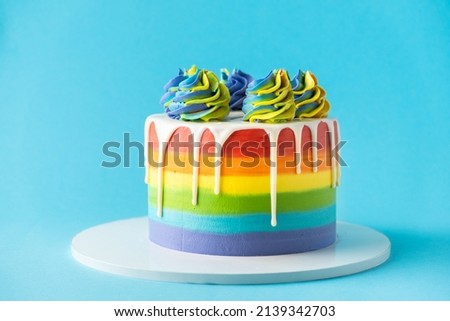 Rainbow cake with whipped cream top on the blue background. Birthday cake with multicolored cream cheese frosting and white chocolate drips.