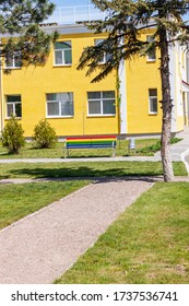 Rainbow colored bench on the background of yellow building facade. Bright colorful street photography, multicolor exterior architecture design. Summer sunny day, empty park with no people.