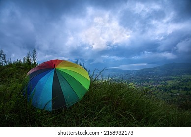 A rainbow colored umbrella enjoying and relaxing on the green hillside against the dramatic blue backdrop of monsoon clouds, beautifully lit green fields and the glistening waters of a lake.    - Shutterstock ID 2158932173
