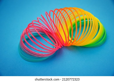 Rainbow Colored Wire Spiral Toy On Blue Background. Toy Plastic Rainbow, Color Spiral For Play. Colorful Plastic Spring Toy Opened To Form An Arc, Coil