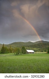 A rainbow in a dark cloudy sky rises above this barn.