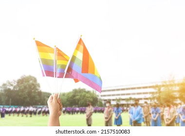 Rainbow Flag, A Symbol For The LGBT Community, Waving In The Wind With Blur Asian Students In The Morning Activities Background. Concept For Supporting And Campaigning The Lgbtq+ Communities At School