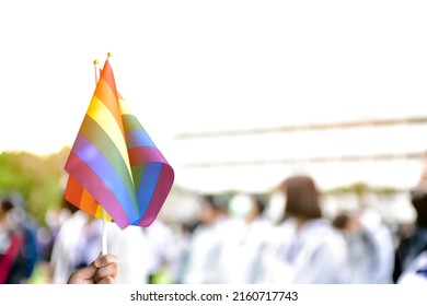 Rainbow Flag, A Symbol For The LGBT Community, Waving In The Wind With Blur Asian Students In The Morning Activities Background. Concept For Supporting And Campaigning The Lgbtq+ Communities At School