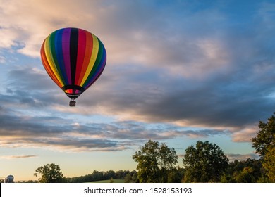 Rainbow Hot-air Balloon Floats Over Grassy Field And Trees At Sunrise